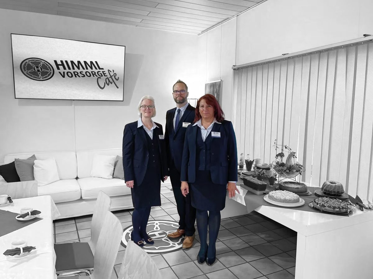 Drei Personen in professioneller Geschäftskleidung stehen bei einer Veranstaltung des HIMML VORSORGE Cafés vor einem Wandschild neben einem Tisch mit Erfrischungen.