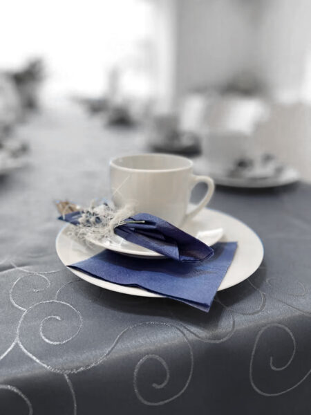 Eine weiße Teetasse mit Untertasse steht auf einem weißen Teller neben einer blauen Serviette und Besteck, alles auf einem Tisch, der mit einer silbernen Tischdecke mit Wirbelmuster bedeckt ist. Der Hintergrund ist sanft unscharf, was die ruhige Atmosphäre vermittelt, die in einem Erfahrungsbericht über das zweite „HIMML VORSORGE Café“ beschrieben wird.
