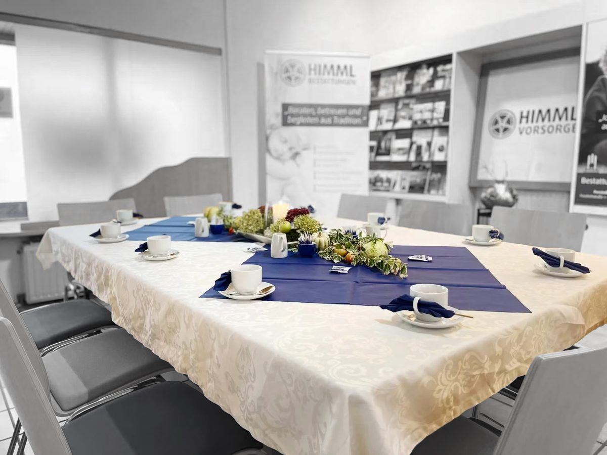 Ein Konferenztisch im HIMML VORSORGE Café, gedeckt mit weißen Kaffeetassen auf blauen Servietten und einem Blumengesteck in der Mitte, in einem Büro, dessen Wände mit dem HIMML-Logo verziert sind.