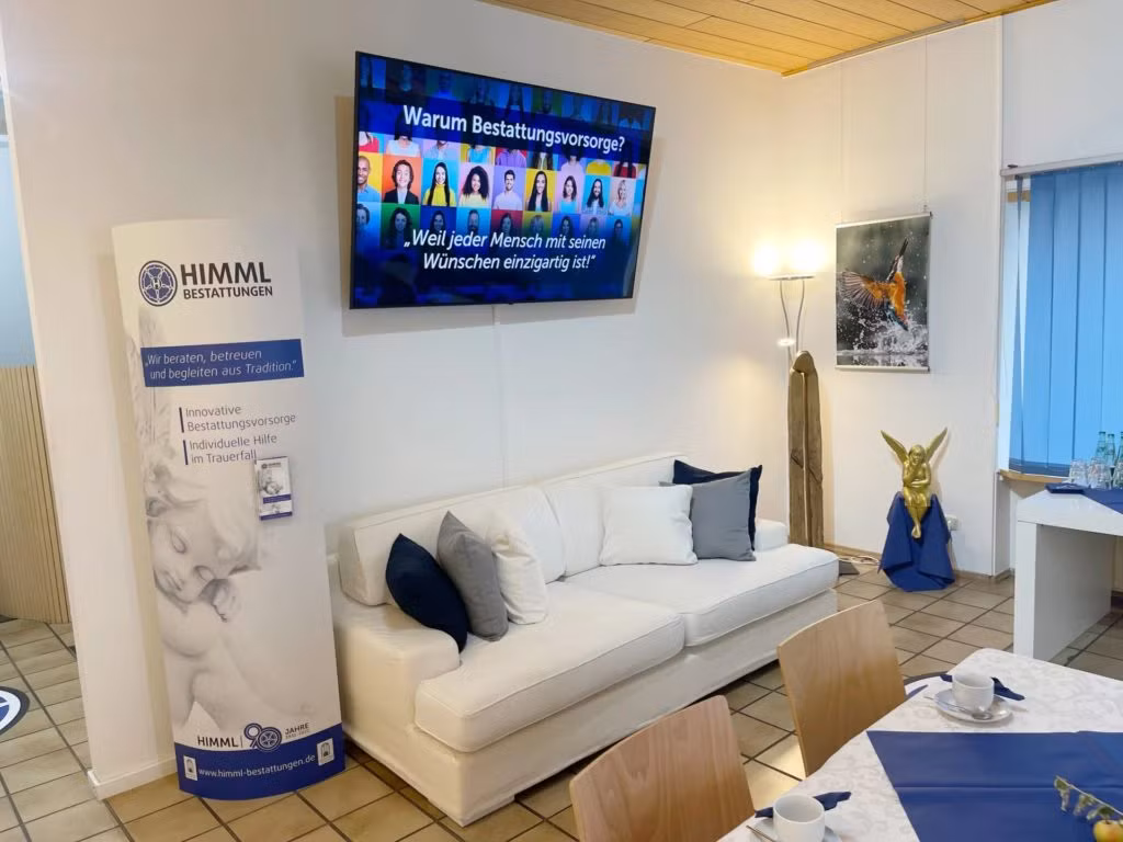 Ein gemütlicher Raum mit einem weißen Sofa, dekorativen Kissen und einem großen Fernsehbildschirm, auf dem Informationen in deutscher Sprache angezeigt werden. Es gibt ein Banner für HIMML Bestattungen, einen gedeckten Tisch für Tee und eine Engelsstatue neben einem kleinen, blau gedeckten Tisch.