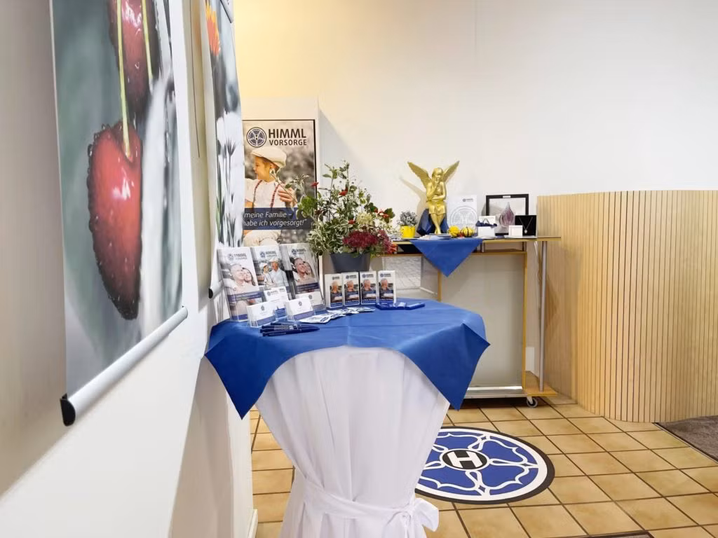 Auf einem kleinen, mit einem blau-weißen Tuch bedeckten Ausstellungstisch liegen Broschüren, Blumen und gerahmte Fotos. Eine goldene Engelsstatue und weitere Blumen stehen auf einem Wagen in einem hellen, gekachelten Raum mit einem HIMML VORSORGE-Poster an der Wand.