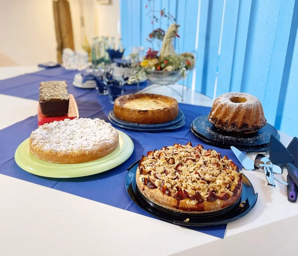 Auf einem Tisch im HIMML VORSORGE Café stehen fünf Torten auf Tellern - mit Früchten belegt, mit Puderzucker bestäubt und im Guss. Teller, Tassen, Besteck und blaue Tischdecken vervollständigen das Ambiente in einem hellen Raum mit blauen Jalousien.