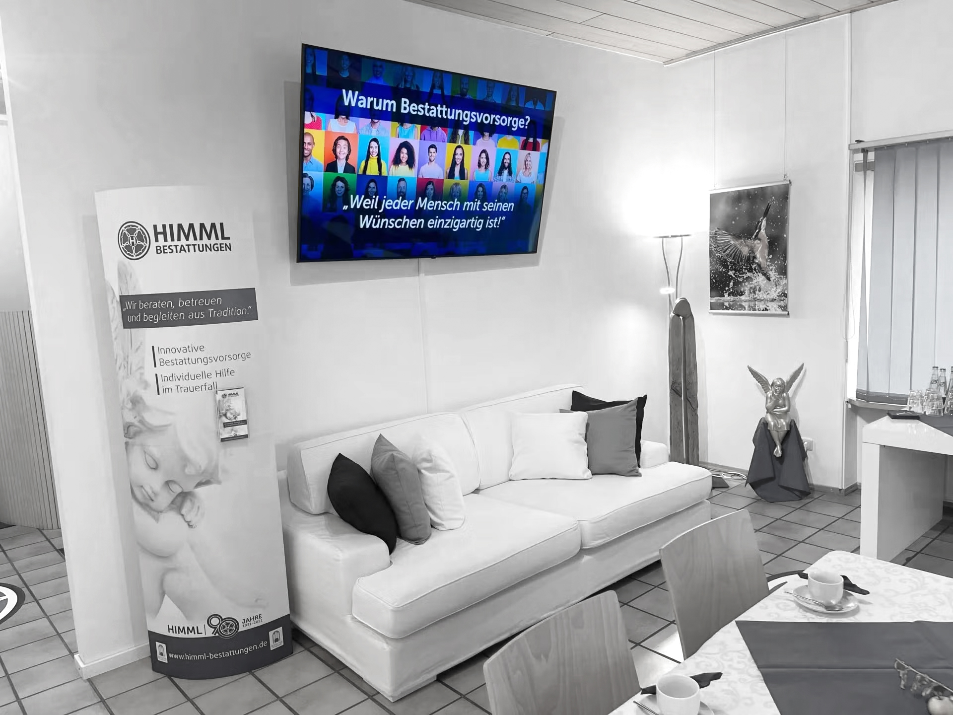 Ein Wartezimmer mit einem weißen Sofa, einem Standbanner und einem Fernseher an der Wand, auf dem eine Präsentation auf Deutsch läuft, lädt zum stillen Nachdenken ein. Außerdem stehen Tische mit Tassen und einer dekorativen Engelsstatue am Fenster, was eine Atmosphäre schafft, die gut zu einer Bestattungsvorsorge passt.