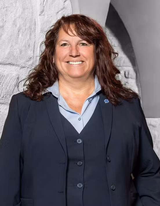 Eine Frau mit welligem braunem Haar lächelt. Sie trägt einen marineblauen Anzug und ein hellblaues Hemd und steht vor einer hellen Steinmauer. Sie verkörpert die Professionalität und Zugänglichkeit, die jedes Teammitglied und jeder Mitarbeiter schätzt.