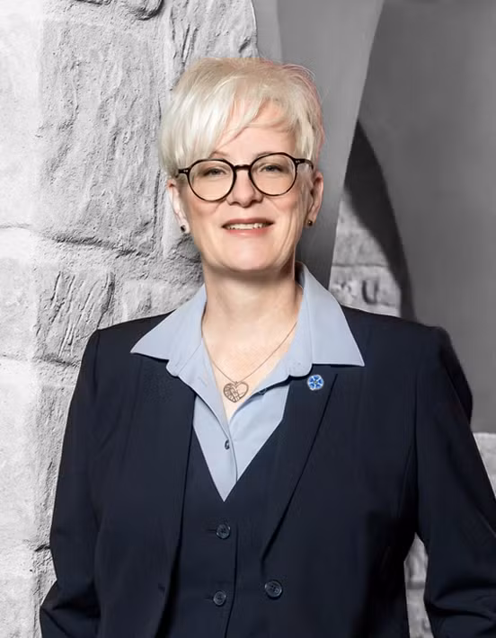 Eine Person mit kurzen weißen Haaren, Brille und einem dunklen Anzug steht vor einer Wand mit Steinstruktur und einem grauen Hintergrund und lächelt – sie verkörpert die Professionalität und Herzlichkeit unseres Teams.