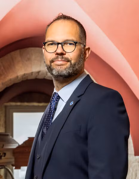 Ein Mann mit kurzen Haaren, gestutztem Bart und Brille trägt einen dunklen Anzug, ein hellblaues Hemd und eine gemusterte Krawatte. Er steht in einer Halle mit roter und steinerner Bogenarchitektur und stellt einen professionellen Mitarbeiter dar, der bereit ist, sein Team zu unterstützen.