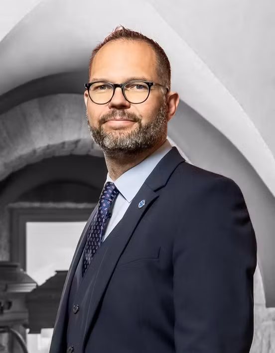 Ein Mann in Anzug und Krawatte, mit Brille und Bart, steht in einem Innenraum vor einem architektonischen Hintergrund mit Bogen und blickt mit einem leichten Lächeln in die Kamera – er ist einer der engagierten Mitarbeiter aus Team 3.