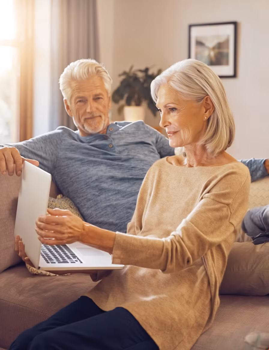 Eine ältere Frau sitzt auf einer Couch und benutzt ihren Laptop, um einen Online-Vorsorgeberater zu konsultieren, während ein älterer Mann neben ihr sitzt und zusieht. Sie wirken entspannt in einem gemütlichen, sonnendurchfluteten Wohnzimmer.