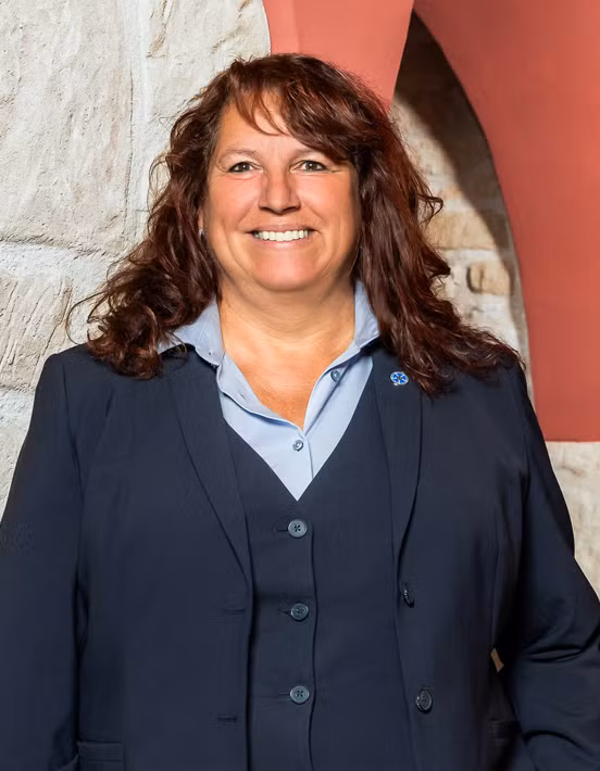 Eine Frau mit gewelltem braunem Haar, die zum Team der Bestattungsvorsorge gehört, trägt einen dunklen Anzug und ein hellblaues Hemd, während sie vor einer Steinmauer und einem roten Bogen lächelt.