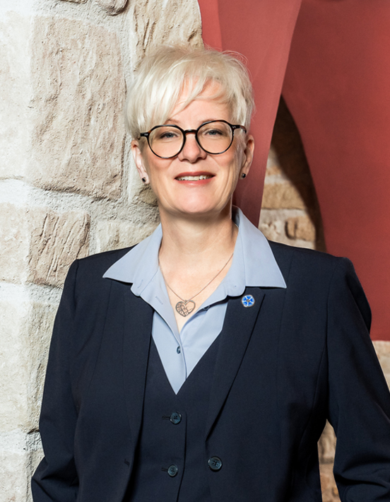 Eine Person mit kurzen weißen Haaren und Brille lächelt, trägt einen marineblauen Anzug und ein hellblaues Hemd und steht in einem Innenraum neben einer strukturierten Steinmauer - Teil des Teams für die Bestattungsvorsorge - mit einem gewölbten Eingang im Hintergrund.