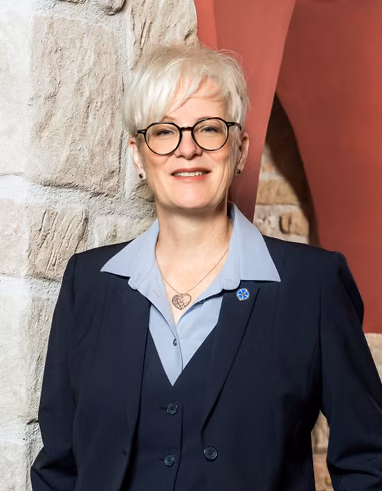 Eine Person mit kurzen weißen Haaren und Brille lächelt, trägt einen marineblauen Anzug und ein hellblaues Hemd und steht in einem Innenraum neben einer strukturierten Steinmauer - Teil des Teams für die Bestattungsvorsorge - mit einem gewölbten Eingang im Hintergrund.