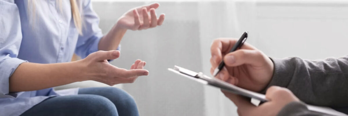 Eine Person gestikuliert mit den Händen, während sie mit einer anderen Person spricht, die auf einem Klemmbrett schreibt, was auf eine Therapie- oder Beratungssitzung hindeutet.