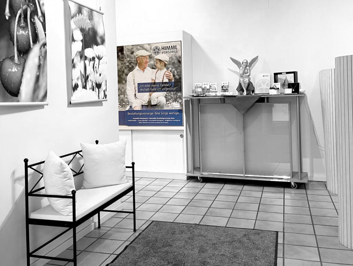 Ein Wartebereich mit einer Bank, Kissen und Fotos an der Wand. In einer Vitrine stehen Engelsfiguren und gerahmte Bilder. Im Hintergrund ist ein Plakat zu sehen, das für Bestattungsdienstleistungen wirbt.