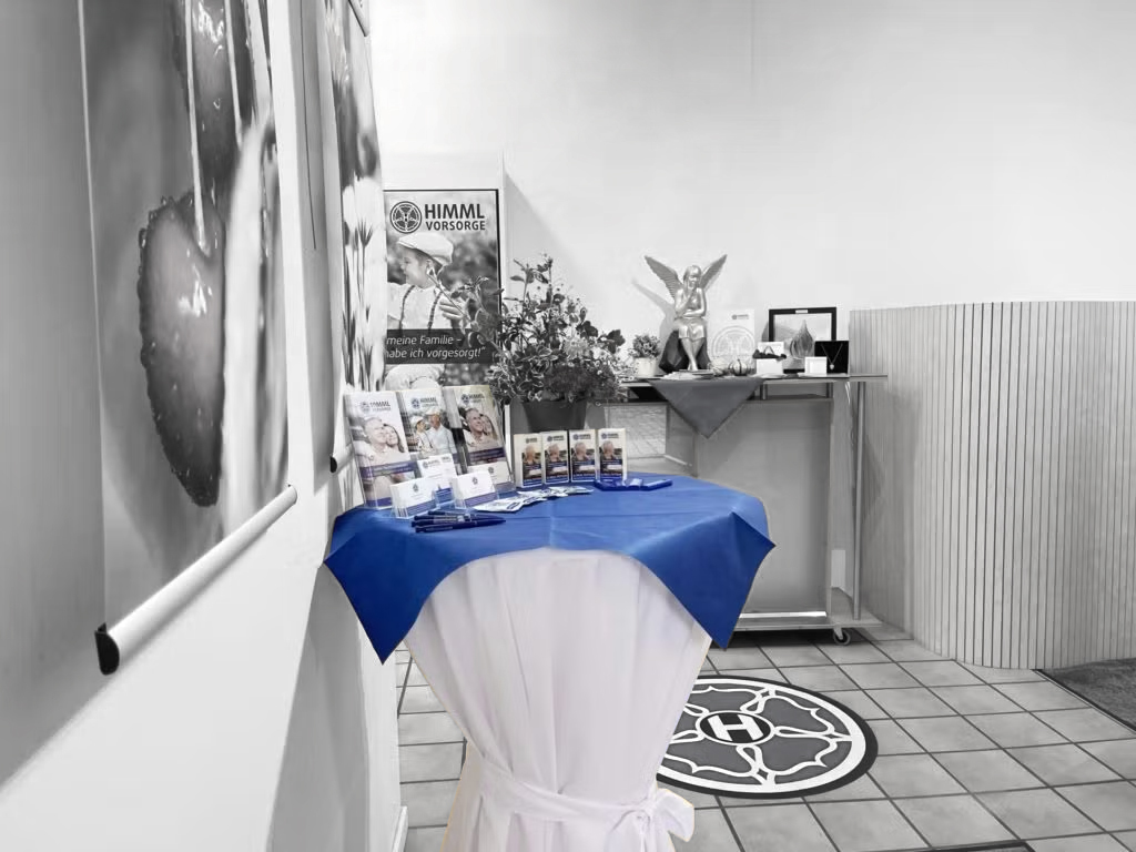 Auf einem mit einem blauen Tuch bedeckten Tisch liegen Broschüren aus. Dahinter befinden sich ein Banner, Blumen und ein Ständer, auf dem eine Statue und ein gerahmtes Foto stehen. Die Kulisse scheint ein Büro oder ein Empfangsbereich mit Fliesenboden zu sein.
