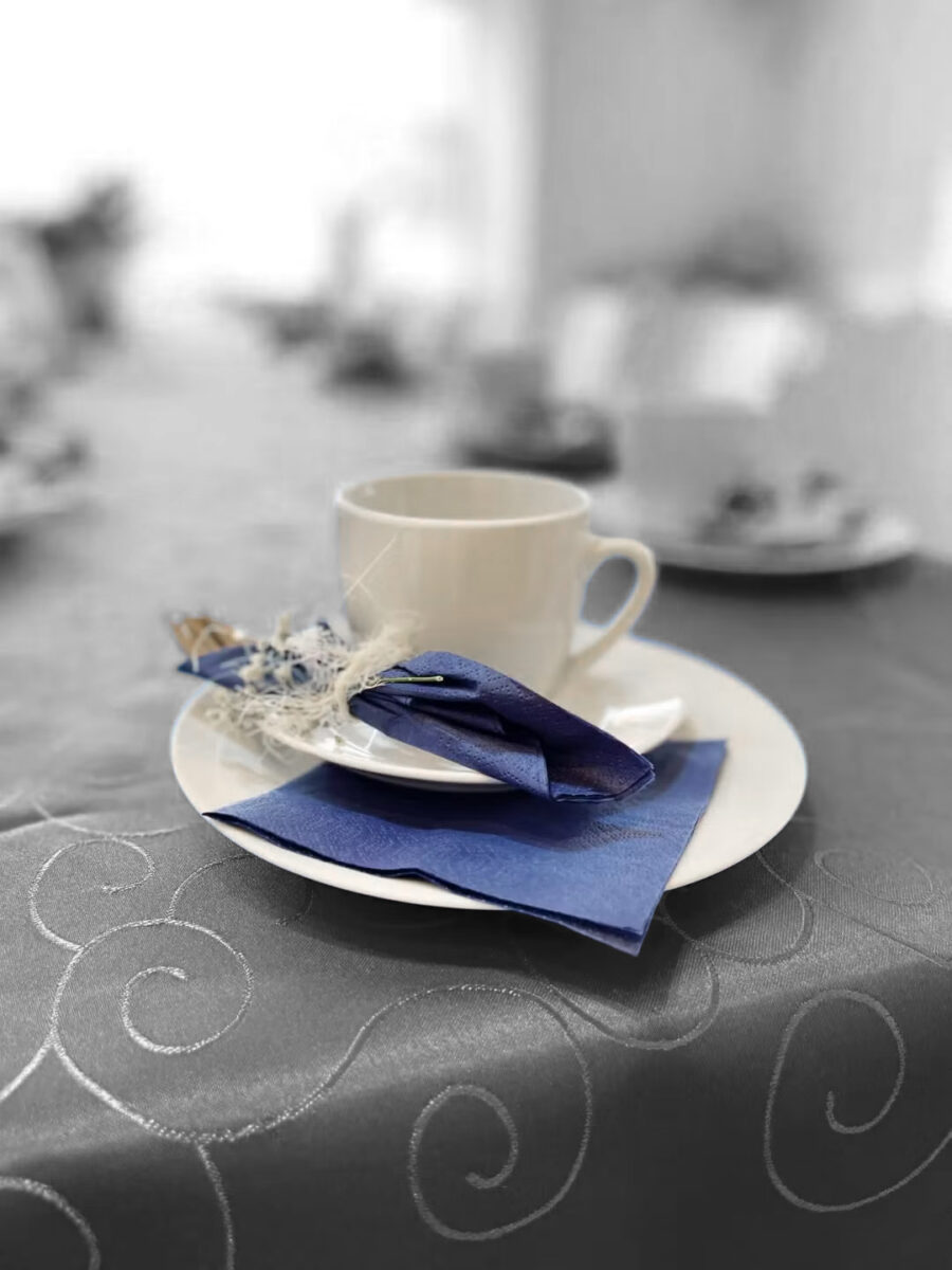 Eine weiße Tasse und Untertasse stehen auf einem weißen Teller, dazu eine blaue Serviette und eine dekorative Blume, angeordnet auf einer grauen Tischdecke mit Wirbelmuster. Der Hintergrund ist unscharf.