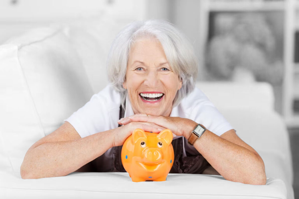 Eine lächelnde ältere Frau mit grauen Haaren lehnt an einem weißen Sofa und stützt ihre Arme auf ein orangefarbenes Sparschwein, was auf Ersparnisse oder finanzielle Sicherheit hindeutet. Sie trägt ein weißes Oberteil und eine Armbanduhr. Der Hintergrund ist sanft unscharf.
