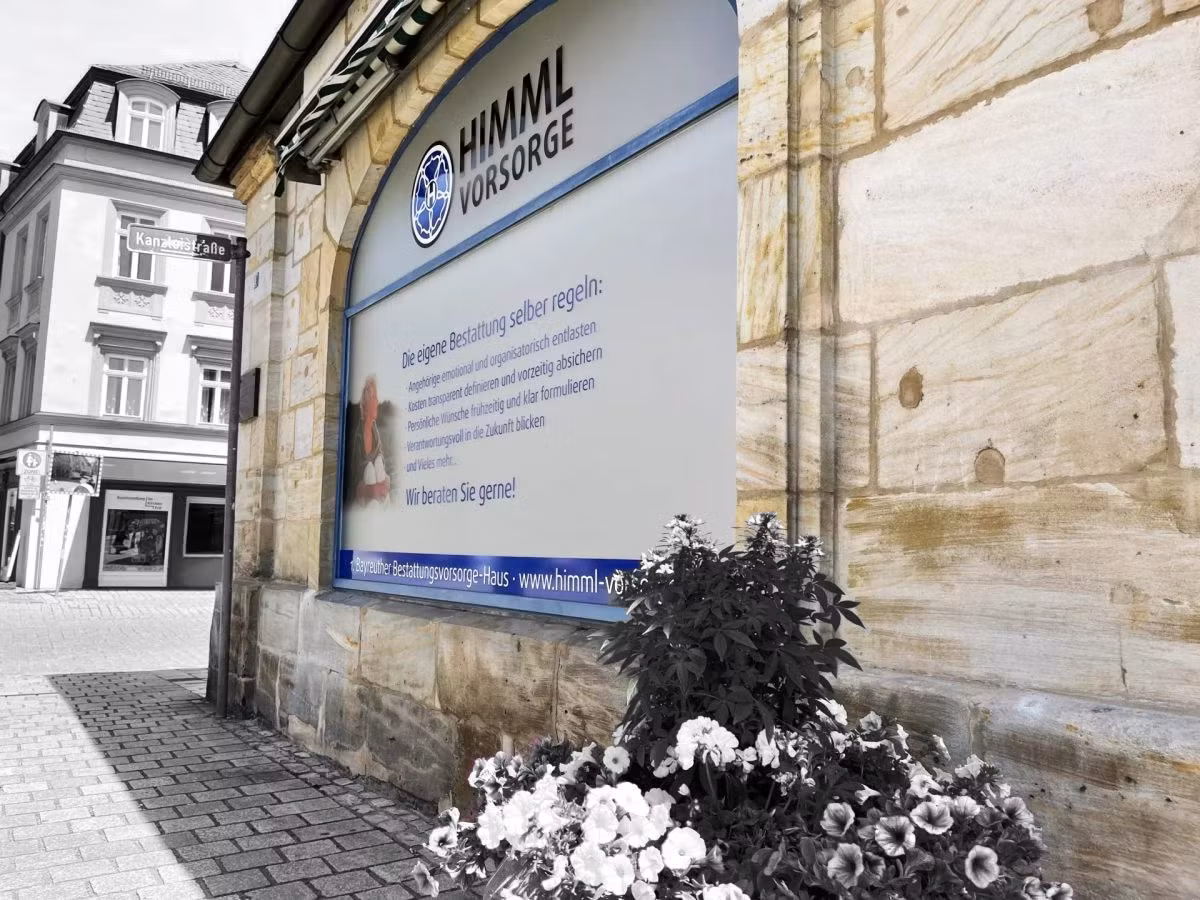 Auf einem Steingebäude prangt ein großes Schild mit der Aufschrift „Himml Vorsorge“, das deutlich hervorsticht. Im Vordergrund sind leuchtende Blumen zu sehen, und im Hintergrund erstreckt sich eine Straße mit Gebäuden. Der größte Teil des Bildes ist schwarz-weiß, abgesehen von den bunten Blumen und einem Teil des „Himml Vorsorge“-Schilds, wodurch ein auffälliger Kontrast entsteht.