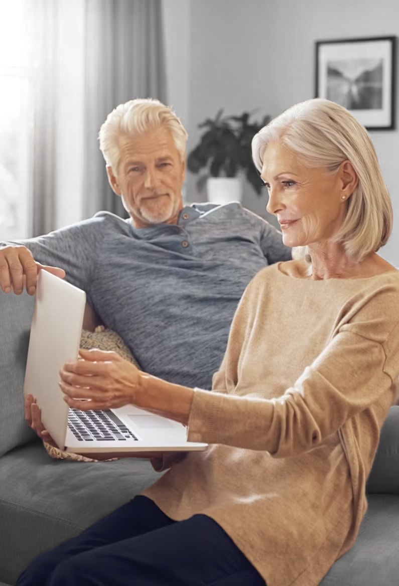 Ein älteres Ehepaar sitzt zusammen auf einem Sofa, lächelt und schaut auf einen Laptop. Die Frau bedient den Laptop, während sich der Mann neben ihr bequem zurücklehnt. Der Raum ist sanft beleuchtet und neutral eingerichtet.