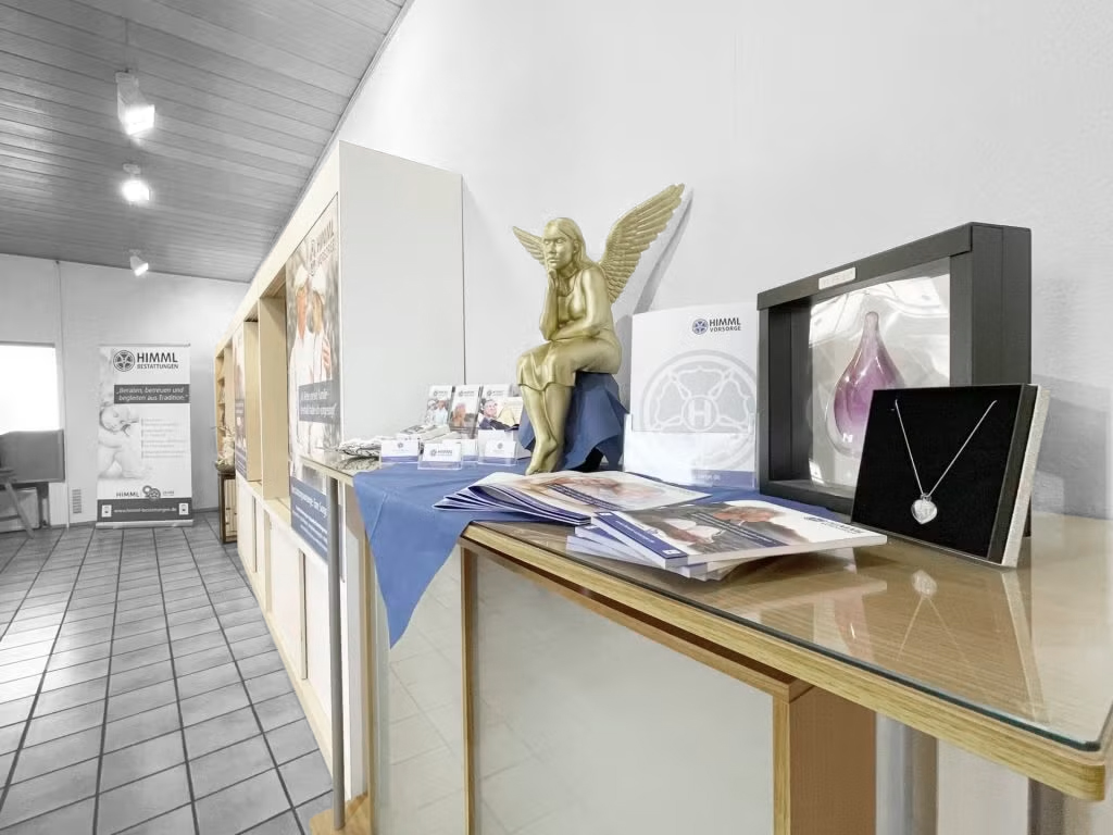 Ein Ausstellungs-Tisch mit Broschüren, einer silbernen Halskette, einem Gedenkobjekt aus Glas und einer goldenen Engelsfigur in einem hell erleuchteten Raum mit Fliesenboden und Informationsplakaten an der Rückwand.
