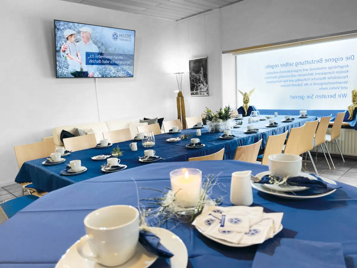 Ein Esszimmer mit Tischen, die für eine Mahlzeit gedeckt sind, mit blauen Tischdecken, weißen Tassen und Tellern, Servietten und Blumengestecken als Tischdekoration. Auf einem Fernsehbildschirm an der Wand werden ein Bild und Text angezeigt. Durch große Fenster fällt Tageslicht herein.