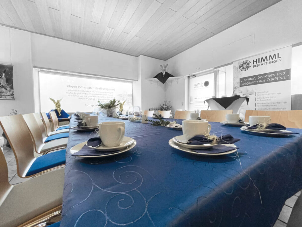 Ein mit Tassen, Tellern und Servietten gedeckter Tisch ist mit einem blauen Tuch bedeckt. Um den Tisch herum stehen Stühle. Im Hintergrund ist ein Schild mit der Aufschrift „HIMML BESTATTUNGEN“ zu sehen, und von der Decke hängen gefaltete Papierkraniche.