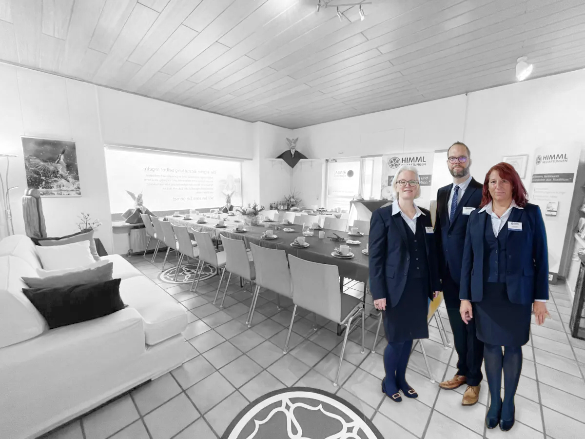 Drei Personen in einheitlicher Geschäftskleidung stehen in einem hellen, modernen Besprechungsraum, in dem ein langer Tisch für ein Essen gedeckt ist. Der Hintergrund ist größtenteils in Schwarz-Weiß gehalten, während die Personen in Farbe dargestellt sind.