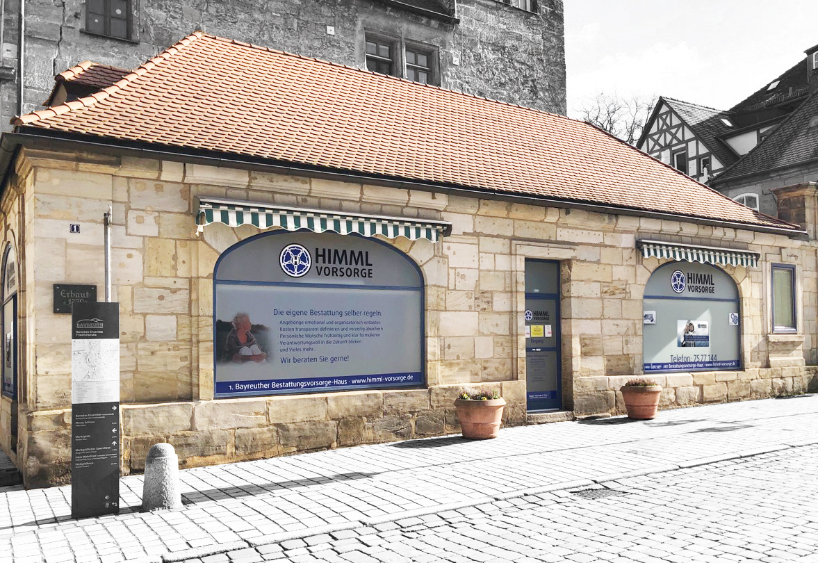 Kleines Steingebäude mit rotem Ziegeldach und großen Fenstern, an denen Schilder mit der Aufschrift „HIMML VORSORGE“ zu sehen sind; dort werden Dienstleistungen zur Bestattungsvorsorge angeboten. Vor dem Gebäude stehen zwei große Topfpflanzen auf einer Kopfsteinpflasterstraße.