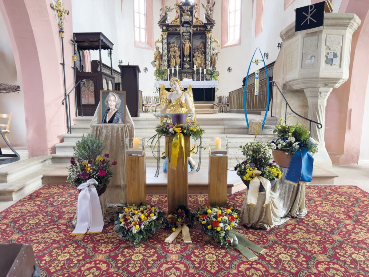 Ein Kirchenaltar mit Blumenkränzen, Kerzen und dem Foto einer Frau, was auf einen Gedenkgottesdienst hindeutet. Der Altar ist mit religiösen Statuen und Dekorationen geschmückt, im Vordergrund liegt ein rot gemusterter Teppich.